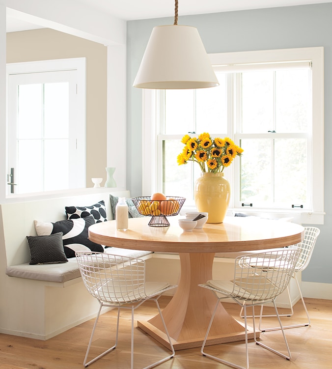 Coin-repas lumineux avec banquettes encastrées, table ronde en bois, chaises blanches en treillis et lampe suspendue blanche.