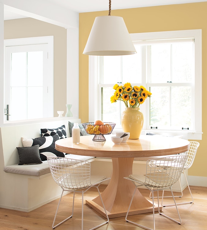 Coin-repas lumineux avec banquettes encastrées, table ronde en bois, chaises blanches en treillis et lampe suspendue blanche.