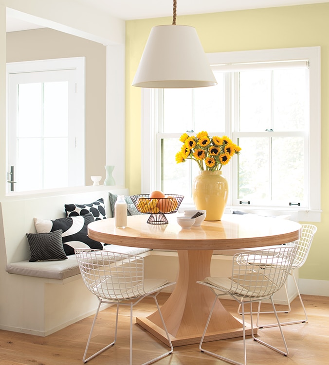 Coin-repas lumineux avec banquettes encastrées, table ronde en bois, chaises blanches en treillis et lampe suspendue blanche.