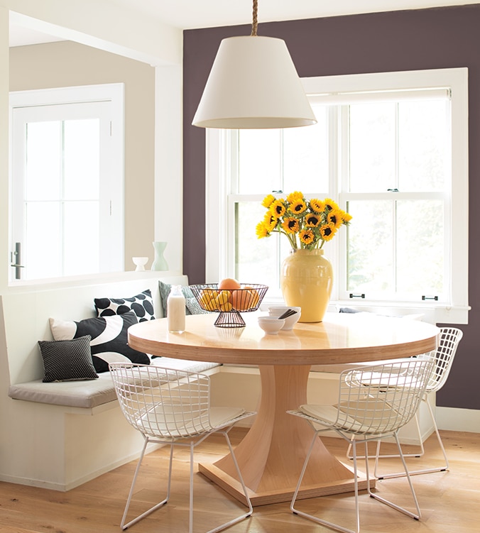 Coin-repas lumineux avec banquettes encastrées, table ronde en bois, chaises blanches en treillis et lampe suspendue blanche.