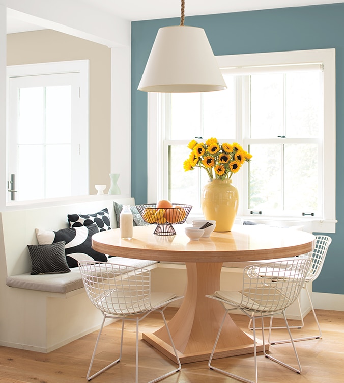 Coin-repas lumineux avec banquettes encastrées, table ronde en bois, chaises blanches en treillis et lampe suspendue blanche.