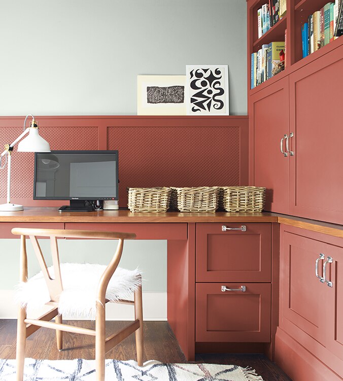 A corner office nook with built-in desk, cabinets and bookshelves, blond wood chair, and white desk lamp.