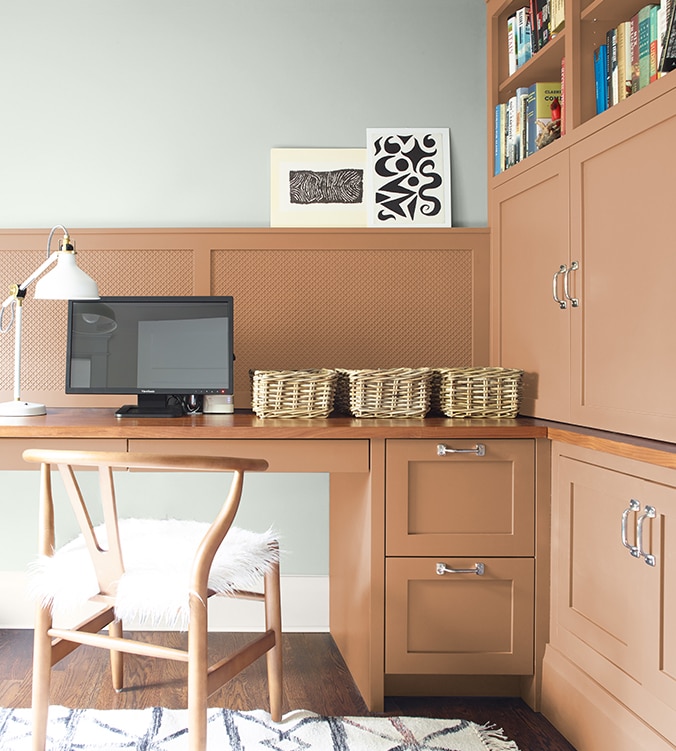 A corner office nook with built-in desk, cabinets and bookshelves, blond wood chair, and white desk lamp.