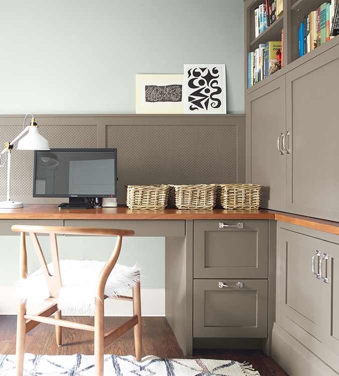 A corner office nook with built-in desk, cabinets and bookshelves, blond wood chair, and white desk lamp.