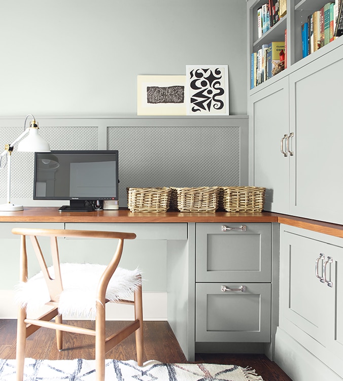 A corner office nook with built-in desk, cabinets and bookshelves, blond wood chair, and white desk lamp.