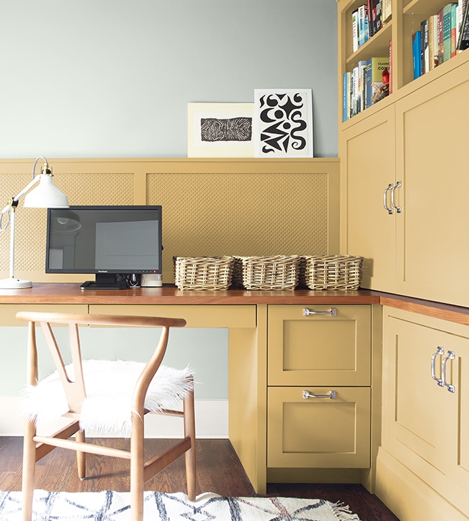 A corner office nook with built-in desk, cabinets and bookshelves, blond wood chair, and white desk lamp.