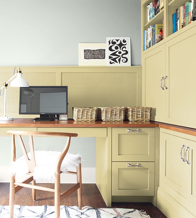 A corner office nook with built-in desk, cabinets and bookshelves, blond wood chair, and white desk lamp.