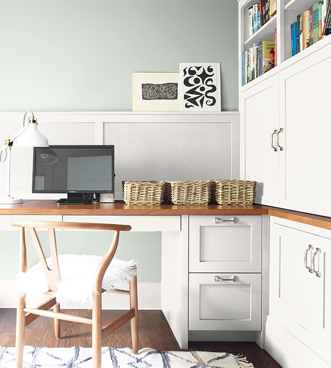 A corner office nook with built-in desk, cabinets and bookshelves, blond wood chair, and white desk lamp.