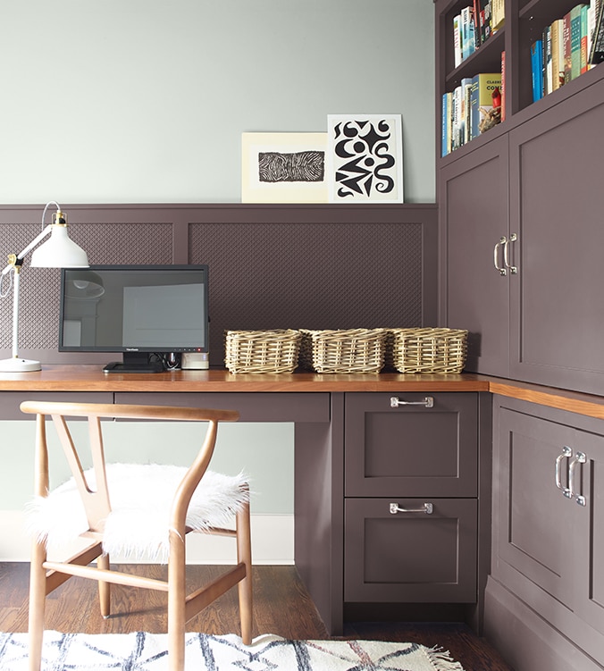 A corner office nook with built-in desk, cabinets and bookshelves, blond wood chair, and white desk lamp.