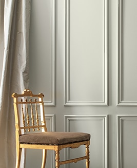 A gray-painted wall and cream-coloured silk curtains frame a formal gold chair.