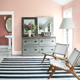 Une chambre à coucher rose au plafond en angle et à la commode grise présente un coin lecture habillé de deux chaises de style contemporain et laisse apercevoir un salon de toilette à l’arrière-plan. Une chambre à coucher rose au plafond en angle et à la commode grise présente un coin lecture habillé de deux chaises de style contemporain et laisse apercevoir un salon de toilette à l’arrière-plan.