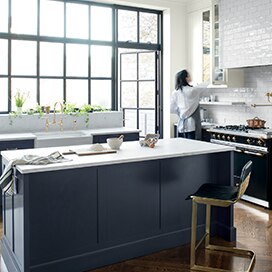 A woman reaches into a white-painted cabinet in an airy kitchen with a navy island. A woman reaches into a white-painted cabinet in an airy kitchen with a navy island.