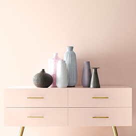 A light pink wall frames a pink console with gold handles and multiple vases. A light pink wall frames a pink console with gold handles and multiple vases.