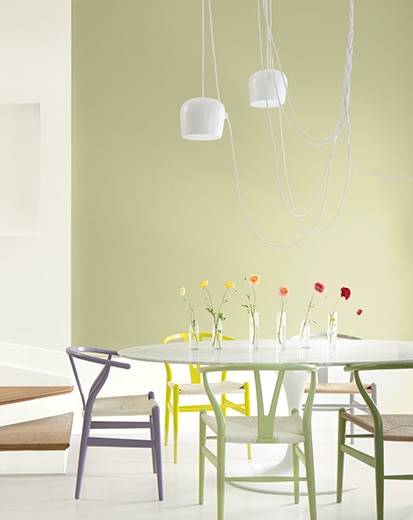 Une salle à manger verte avec des chaises contemporaines multicolores, un luminaire suspendu et plusieurs vases de fleurs posés sur la table.