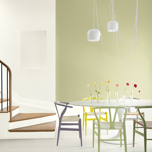 Une salle à manger verte avec des chaises contemporaines multicolores, un luminaire suspendu et plusieurs vases de fleurs posés sur la table.