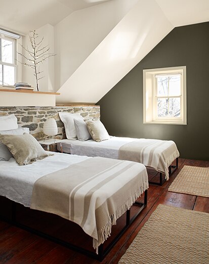White bedroom with dark green-painted accent wall, two beds in front of a stone half wall, hardwood floors, and small window.