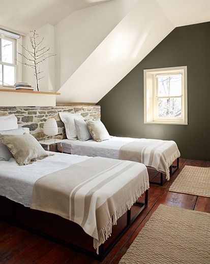 Chambre à coucher blanche avec mur d'accent vert foncé, deux lits devant un demi-mur en pierre, plancher de bois franc et petite fenêtre.