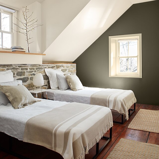 White bedroom with dark green-painted accent wall, two beds in front of a stone half wall, hardwood floors, and small window.