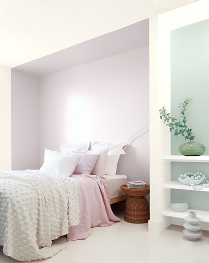 Una habitación blanca con una parte empotrada en violeta y un acento en verde azulado detrás de estantes blancos, cama con ropa de cama blanca y violeta.