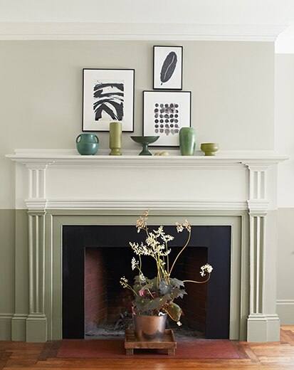 Two-toned fireplace in off white and sage green, with flowering plant in a planter in front of the hearth; art on walls.