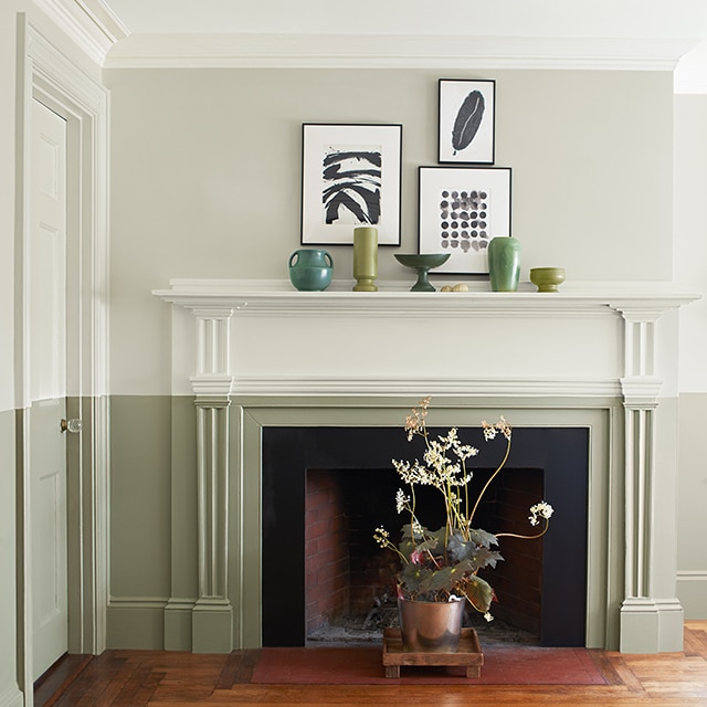 Two-toned fireplace in off white and sage green, with flowering plant in a planter in front of the hearth; art on walls.