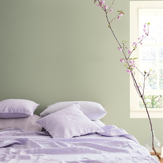Une chambre à coucher avec literie lilas, mur vert sauge et arbre fleuri en pot.