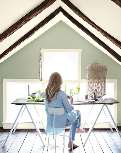 A woman at a desk in a home office with vaulted white ceiling and trim, wooden rafters, large window, and sage-green wall. 