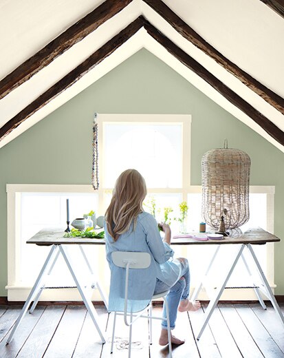 Una mujer en un escritorio en una oficina en casa con techo blanco abovedado y marco, vigas de techo de madera, ventana grande y pared verde salvia.