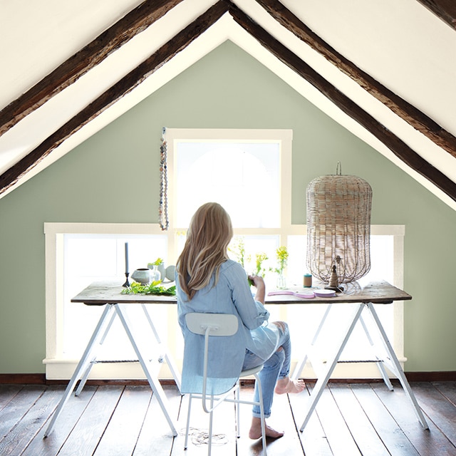 A woman at a desk in a home office with vaulted white ceiling and trim, wooden rafters, large window, and sage-green wall. 