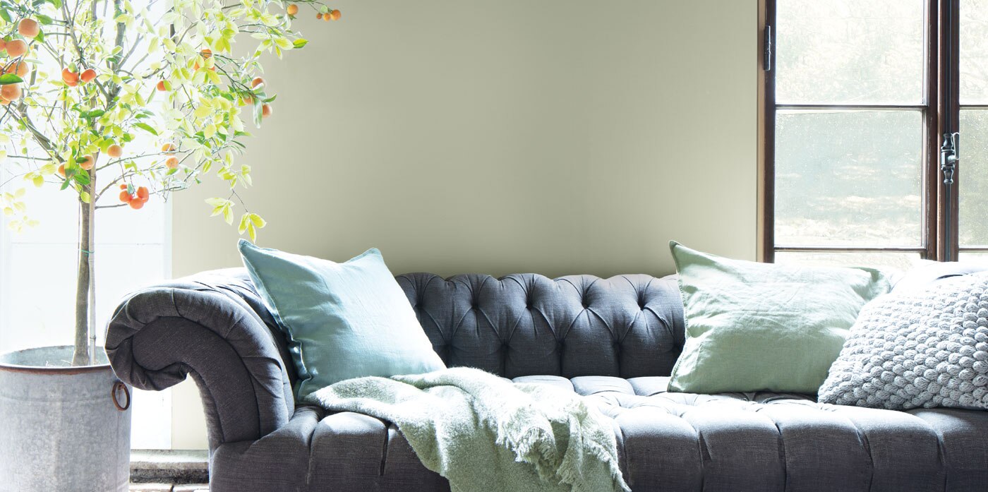 Salon avec un canapé profond de couleur grise, des coussins verts, un arbre en pot et des murs vert sauge avec des moulures en bois.