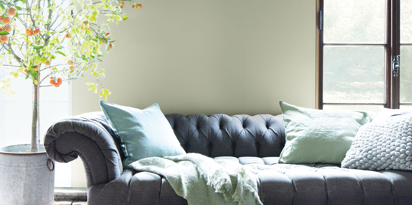 A living room with classic gray couch, green accent pillows, indoor tree, and sage green-painted walls with wood trim.