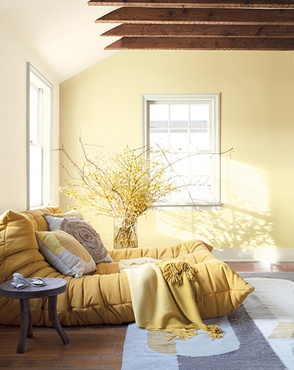 White room with yellow-painted accent wall, wooden rafters, soft yellow chaise, throw pillows, plants, and round end table.