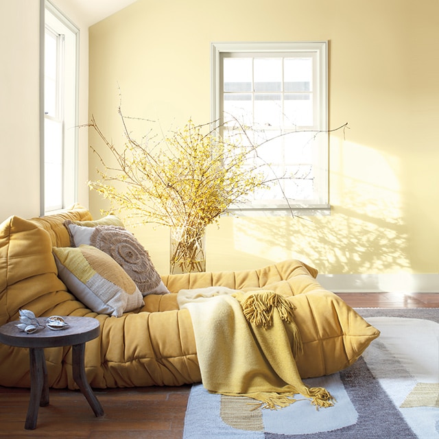 White room with yellow-painted accent wall, wooden rafters, soft yellow chaise, throw pillows, plants, and round end table.