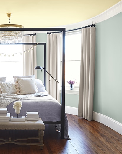 Blue gray-painted bedroom with soft yellow ceiling, large iron-framed bed, small chandelier, and side table with décor.