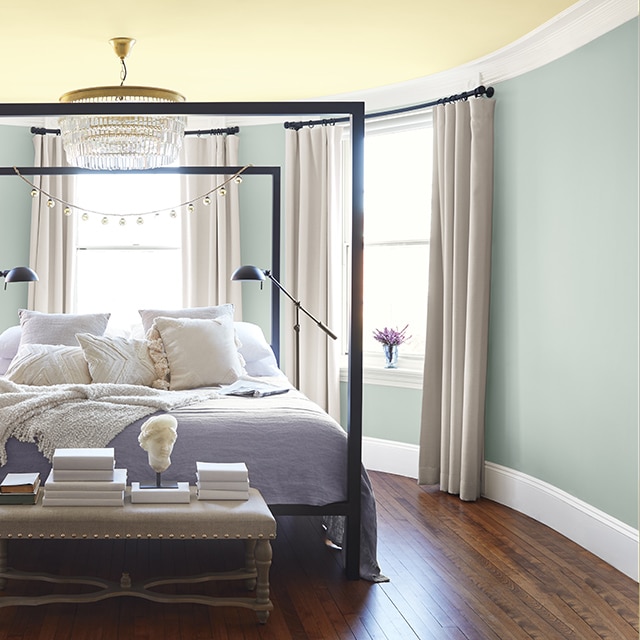 Chambre à coucher bleu-gris avec plafond jaune pâle, grand lit à armature en fer, petit lustre et table d'appui avec objets décoratifs.