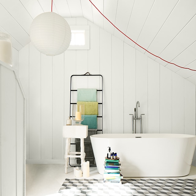 Une salle de bains blanche avec une suspension sphérique, une grande baignoire, des étagères noires, un plafond en planches à feuillure et un tabouret-escabeau blanc.