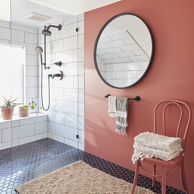 Salle de bains rouge terre avec douche en carreaux blancs, miroir rond, chaise avec serviettes, porte-serviettes et petit tapis beige.