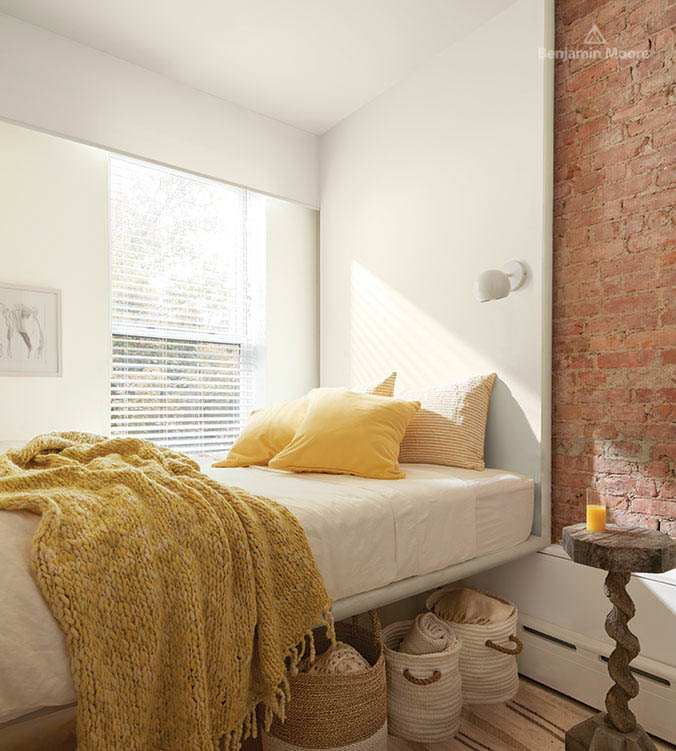 A bedroom with brick wall, wooden side table, white bed, yellow pillows and blankets, and baskets under the bed.