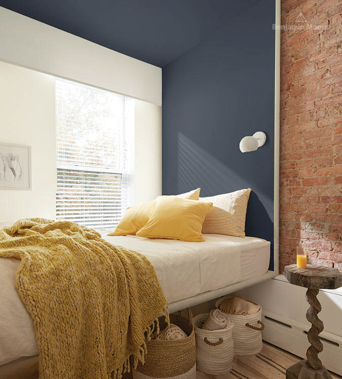 Chambre à coucher avec mur en briques, table d'appui en bois, lit blanc, oreillers et couverture jaunes et paniers entreposés sous le lit.