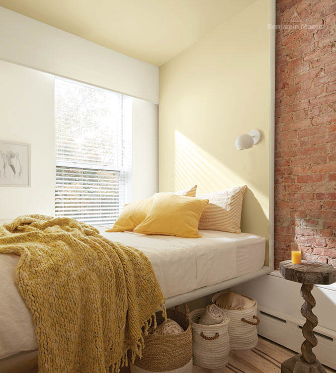 A bedroom with brick wall, wooden side table, white bed, yellow pillows and blankets, and baskets under the bed.