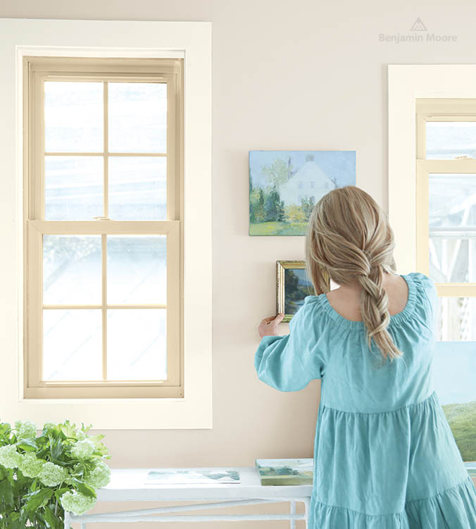 Une femme suspend des tableaux au mur derrière une table blanche dans un bureau à domicile où se trouvent des plantes.