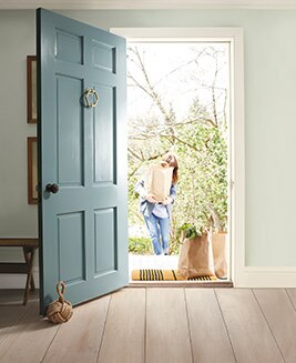 Une porte bleue ouverte dans une pièce blanche laisse voir une propriétaire qui revient avec ses courses. 