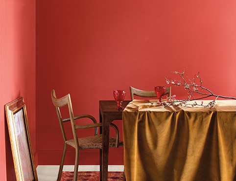 Red orange dining room painted in Raspberry Blush 2008-30 with wooden table, gold tablecloth, tree branches, and a golden frame propped against the wall.