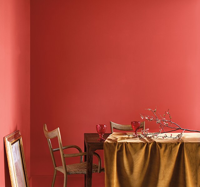 Salle à manger enduite du rouge orangé Tourbillon de Framboises 2008-30 avec table en bois, branche d’arbre posée sur nappe dorée et tableau au cadre doré appuyé contre le mur.