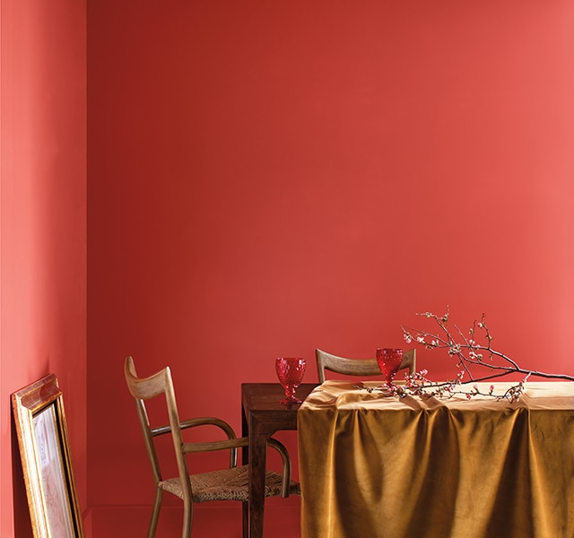 Salle à manger enduite du rouge orangé Tourbillon de Framboises 2008-30 avec table en bois, branche d’arbre posée sur nappe dorée et tableau au cadre doré appuyé contre le mur.