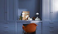 A modern red office chair sits in a home office painted in Blue Nova CC-860, with built-in shelving, cabinetry and in-set desk topped with a pegboard, plants, books and a modern domed white desk lamp.