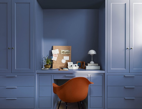 Une chaise moderne rouge dans un bureau peint en Abysse CC-860 avec étagères, armoires et table de travail encastrées, panneau d’accrochage, plantes, livres et lampe de bureau blanche en forme de dôme.