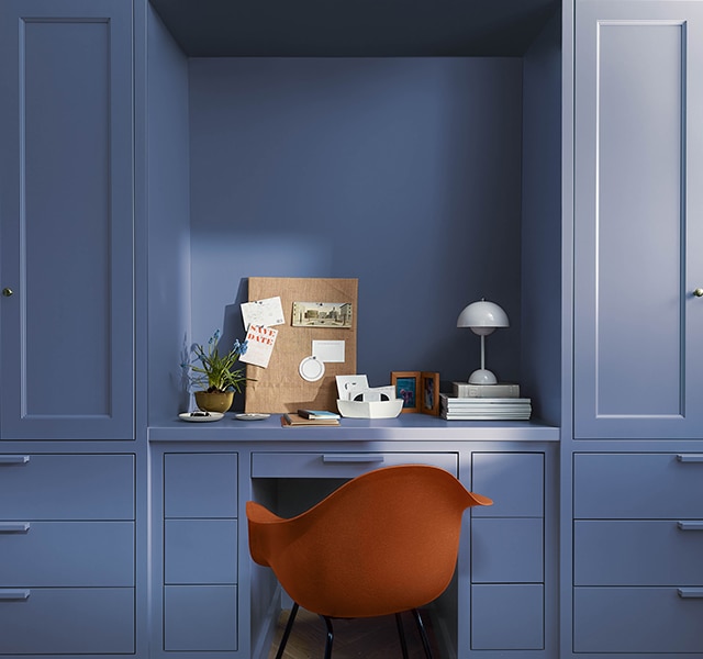 A modern red office chair sits in a home office painted in Blue Nova 825, with built-in shelving, cabinetry and in-set desk topped with a pegboard, plants, books and a modern domed white desk lamp.