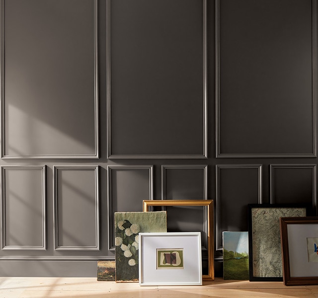 Artwork and frames arranged on a floor against a deep chocolate brown-painted wall with decorative molding. Artwork and frames arranged on a floor against a deep chocolate brown-painted wall with decorative molding.