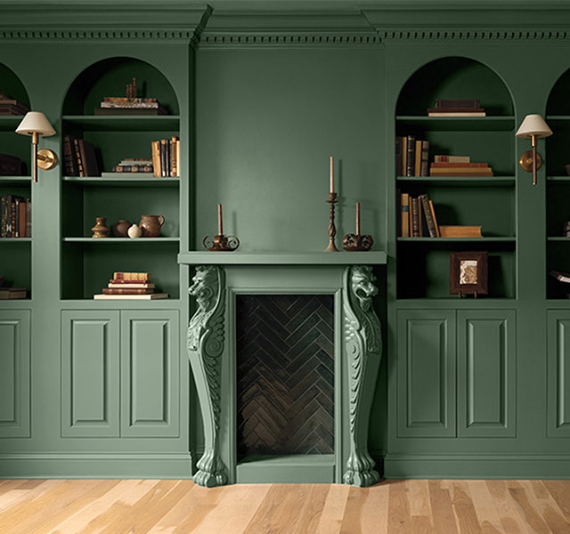 A green painted room with lower panelled walls and arched, built-in shelving topped with books and decor.